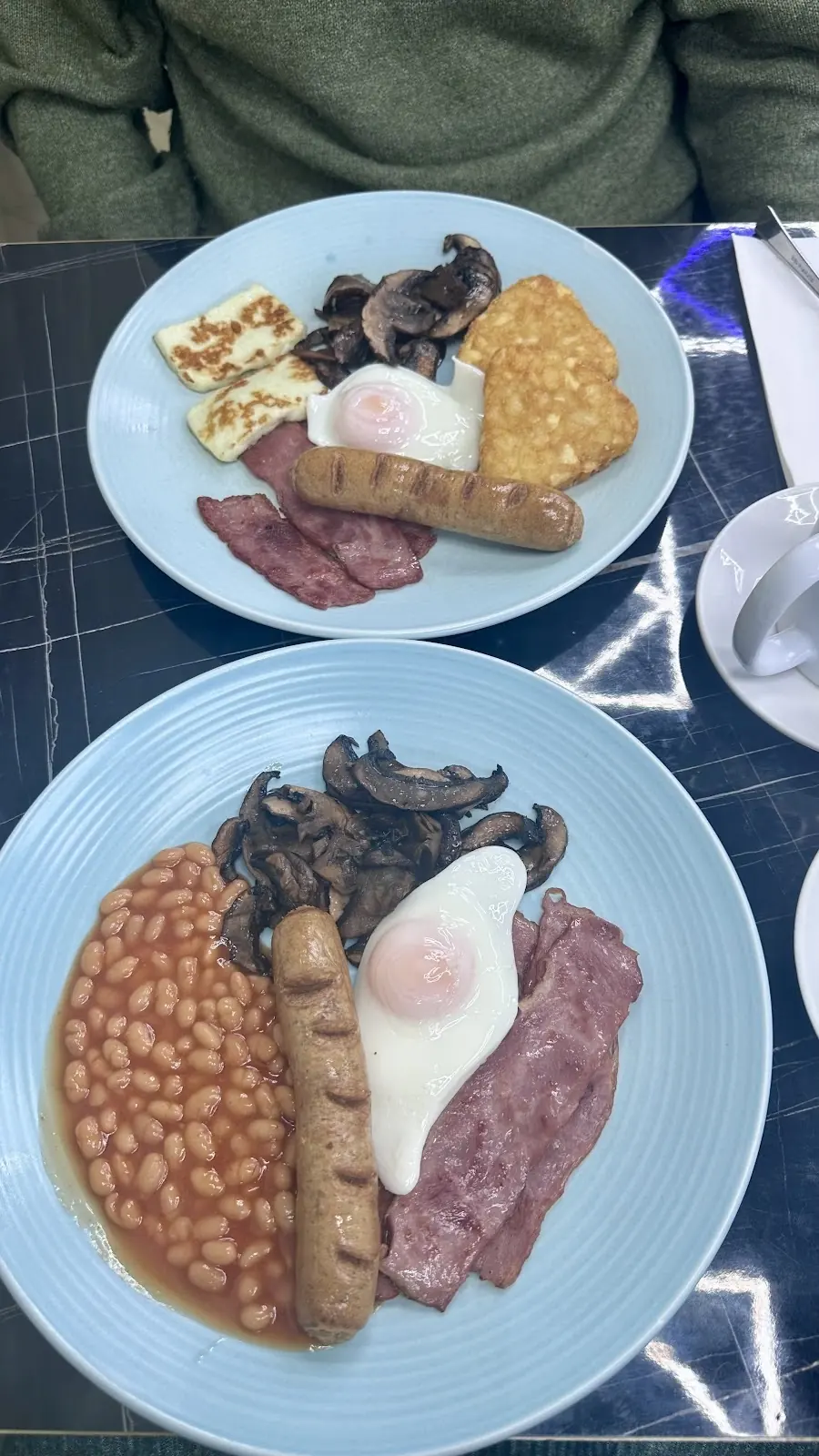 Two breakfast plates side by side on a marble table, one with halloumi, egg and bacon, the other a cooked breakfast