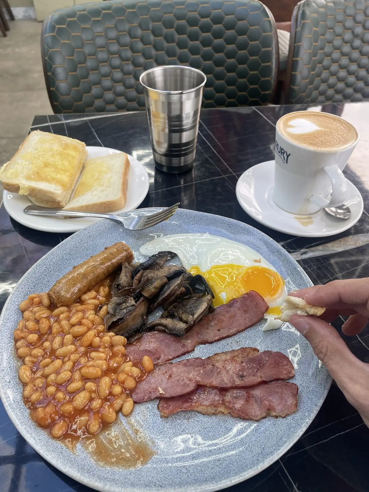 A breakfast plate of beans, bacon, mushrooms and egg on a cafe table, with a latte, toast and cafe chairs behind