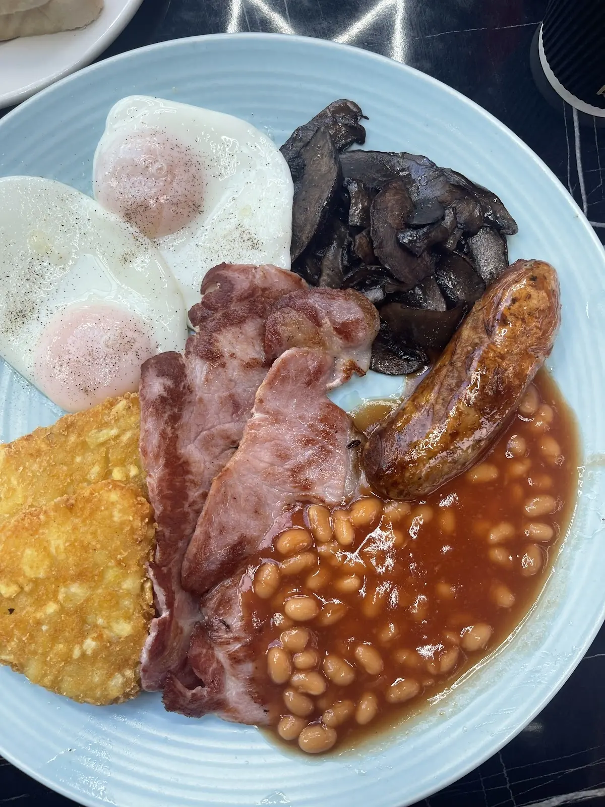 A close-up blue plate of fried eggs, sausages, bacon, mushrooms, hash brown and beans