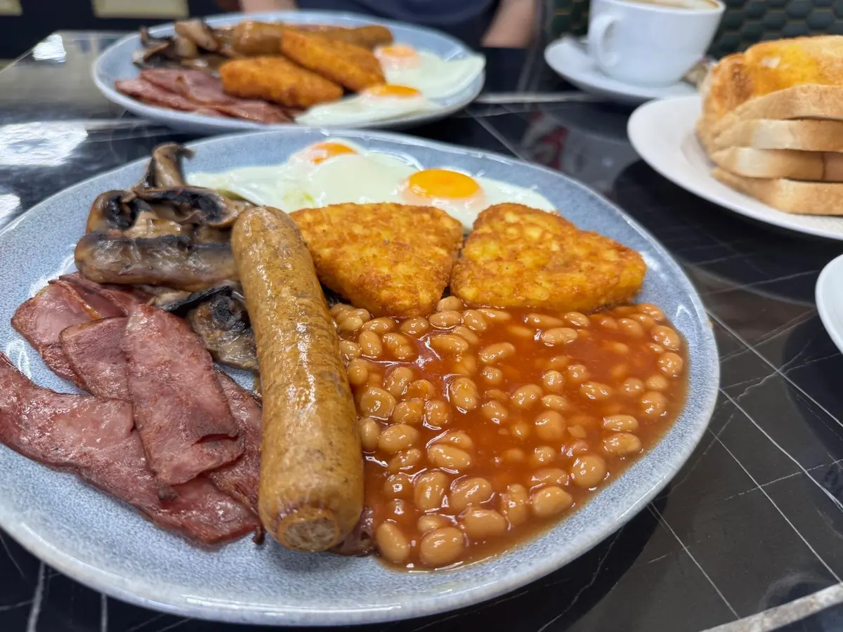 A full English breakfast on a pale blue plate with bacon, sausage, hash brown, mushrooms, fried egg and beans, toast and coffee behind