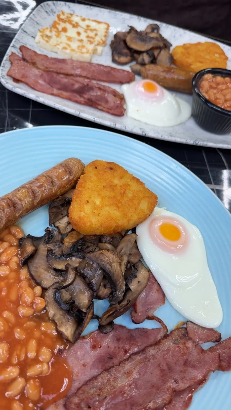 Two cooked breakfast plates shot from above with bacon, mushrooms, hash brown, fried egg and beans