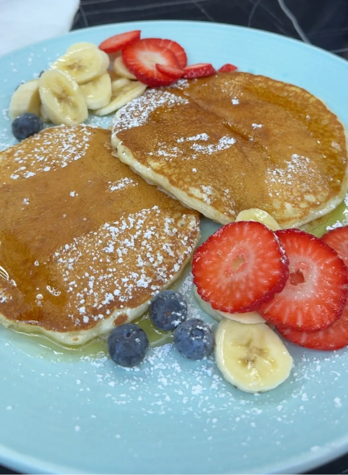 A stack of pancakes dusted with icing sugar, ringed with strawberries, banana and blueberries on a blue plate
